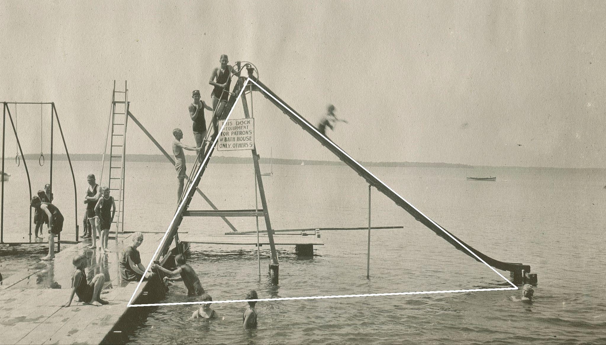 Black & white photo depicting a large slide ending in a lake, with many bathers awaiting their turn to go down. A white triangle traces the shape of the slide.