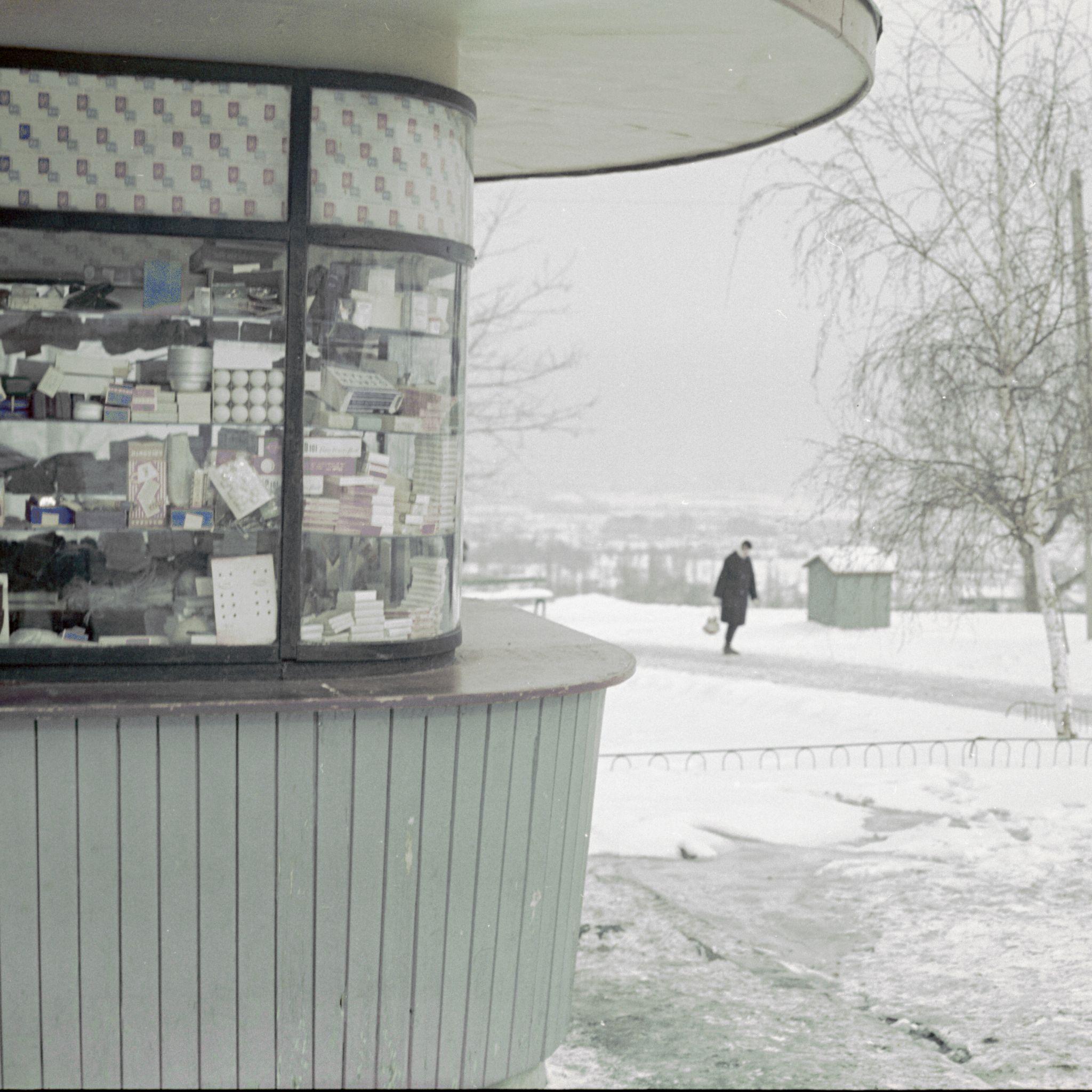 A black and white version of the previous image (a man in a dark coat walking outside in the snow, with a shop in the foreground).