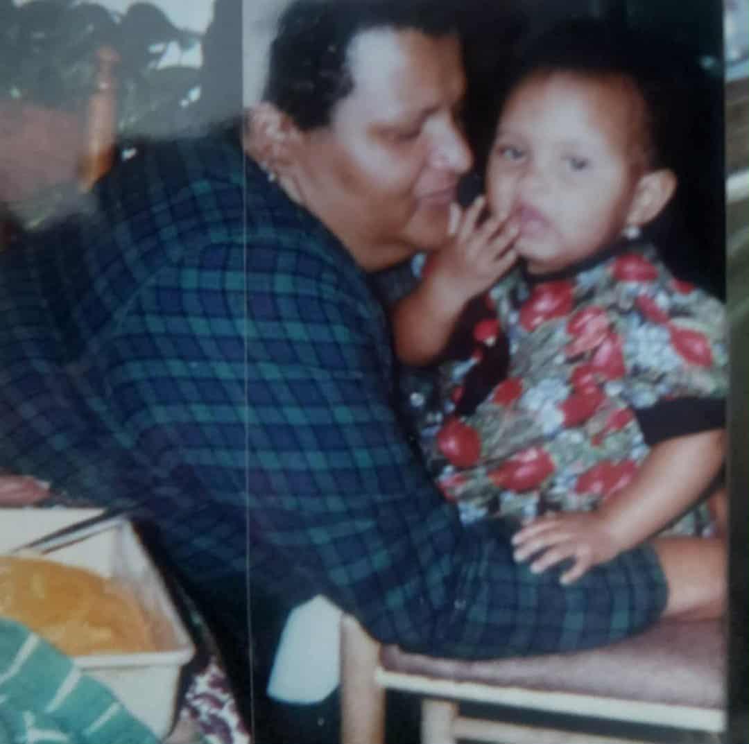 A photo of a young Odassey, around two years old, sitting on a stool while her grandmother, Nana, gently wraps her arms around her to hold her steady. Nana’s face is close to Odassey’s, full of tenderness and quiet pride. On the table beside them, there’s a dish of food, symbolizing the family warmth that surrounded every meal. The lighting is soft and homey, capturing an intimate moment of love, care, and connection between generations.