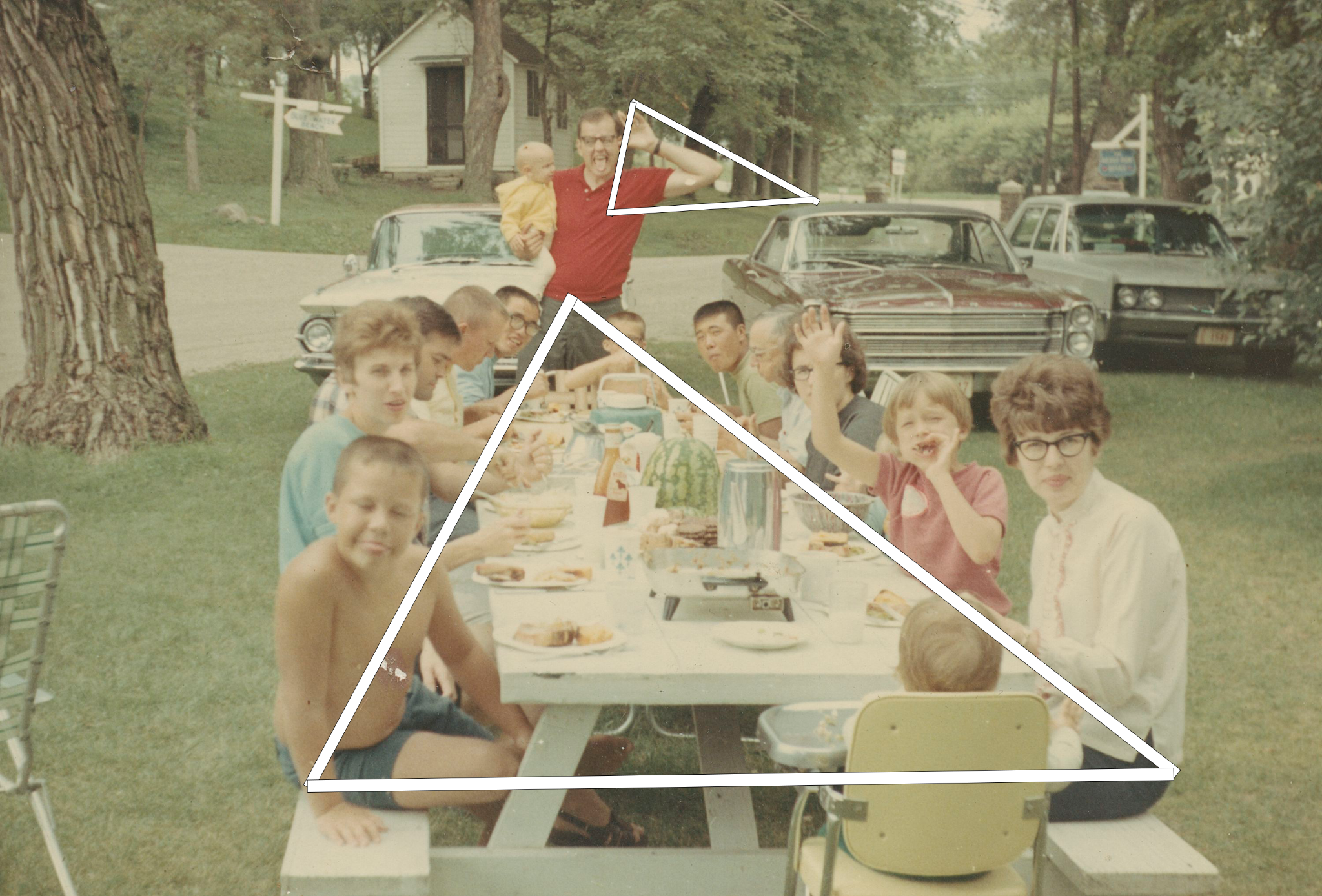 A color photo depicting a long picnic table with many individuals, adults &amp; children. eating a meal. A white triangle connects the sides of the table and another connects a man's outstretched elbow to a car nearby".