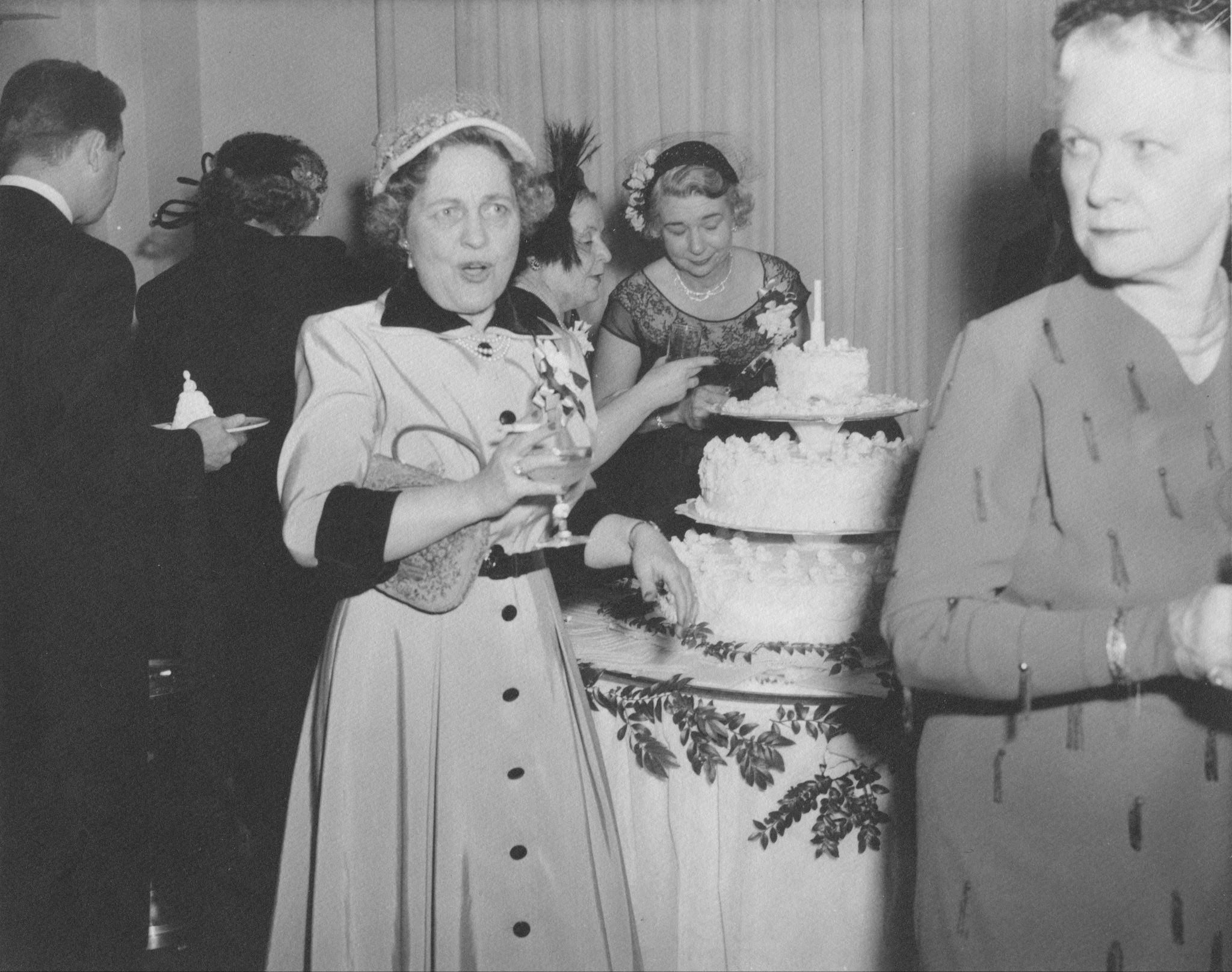A well-dressed woman stands in front of a cake with her wineglass and cigarette in her right hand. There are people all around her looking towards the cake. 
