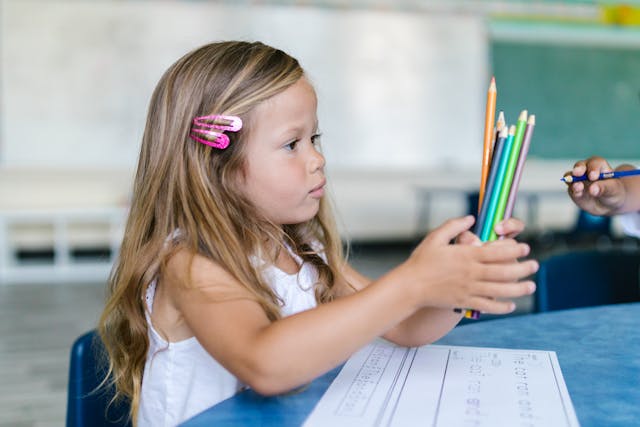 A girl holds 7 colored pencils in both hands. A worksheet sits on the table in front of her.