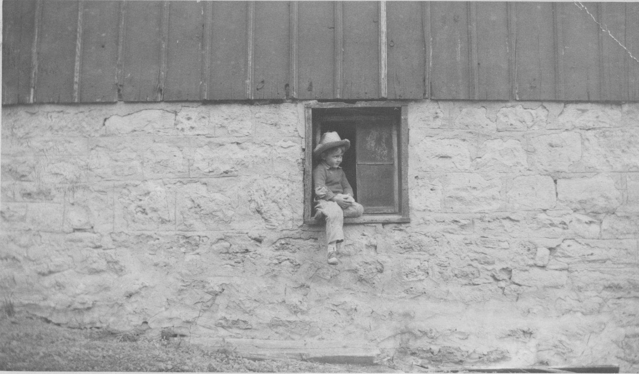A little boy in a cowboy hat sits hanging out of the window of a stone building. 
