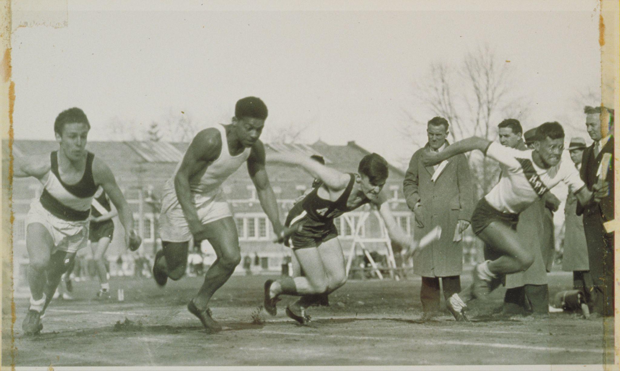 A track race with 4 competitors leaning forward as they run, with several spectators looking on.