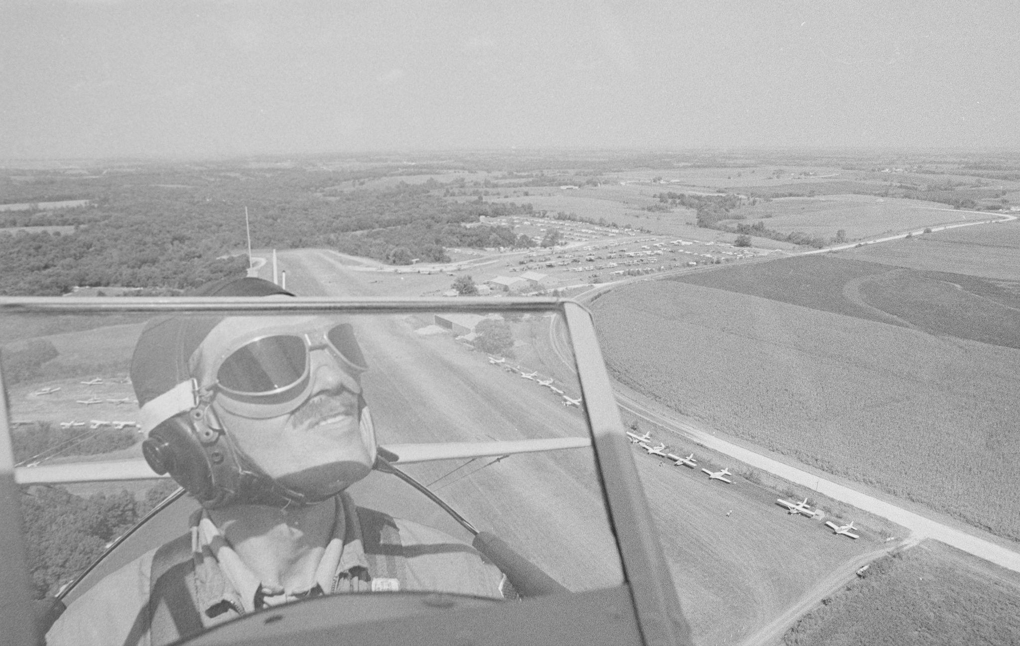 A black & white photo of a pilot wearing helmet and goggles in an open-air cockpit, frying over an airport runway and surrounding area with trees and fields.