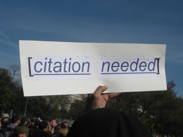 Hand held sign reading “citation needed.”
