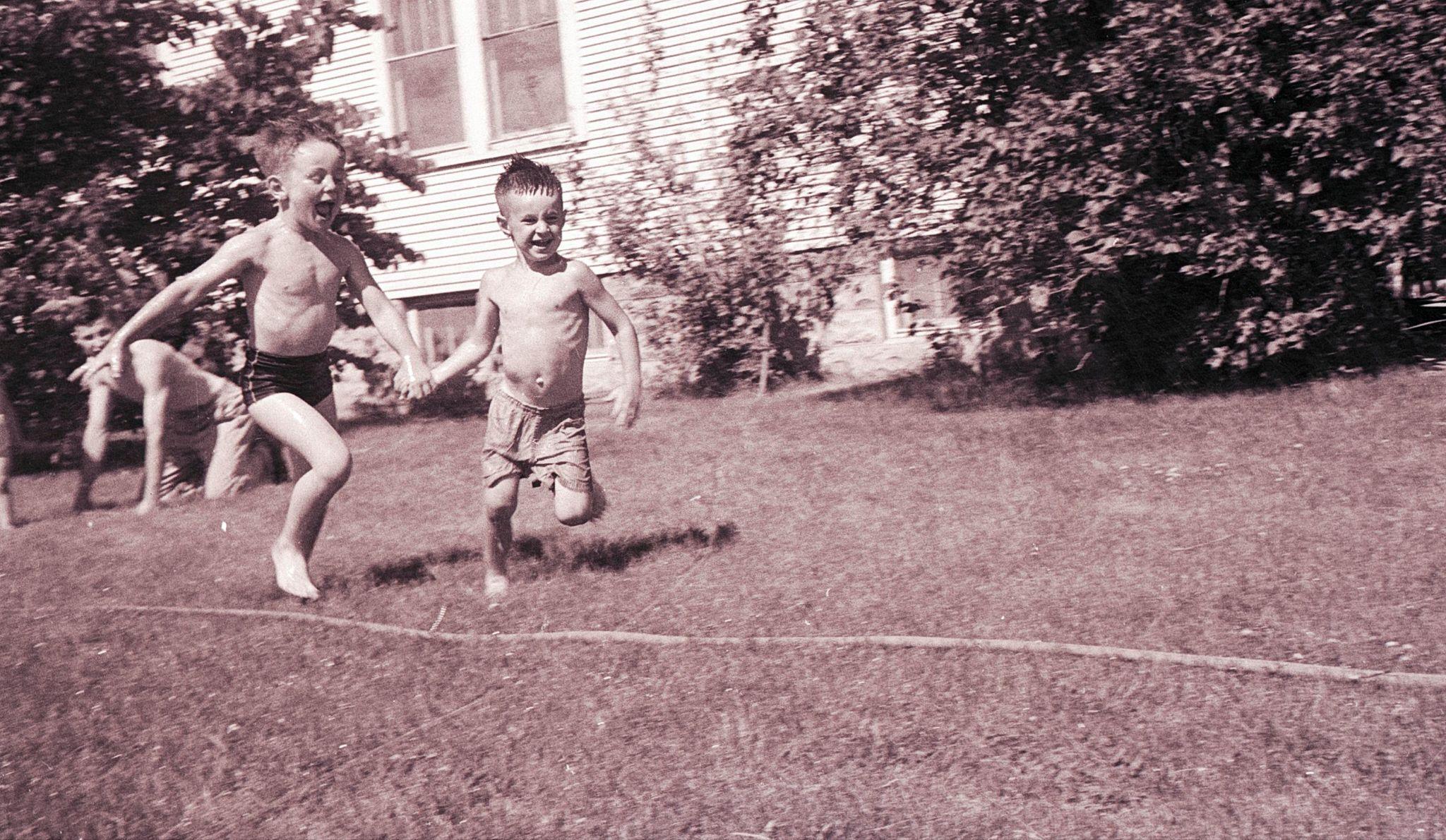 Two boys in swimsuits run happily towards a sprinkler to the right of the photo with a man sitting and laughing behind them. 
