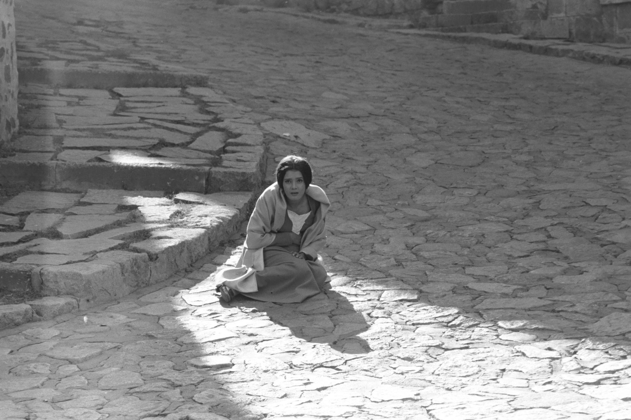 A woman with short hair wearing layered clothing sitting on a cobblestone street near stone steps, with sunlight casting shadows.