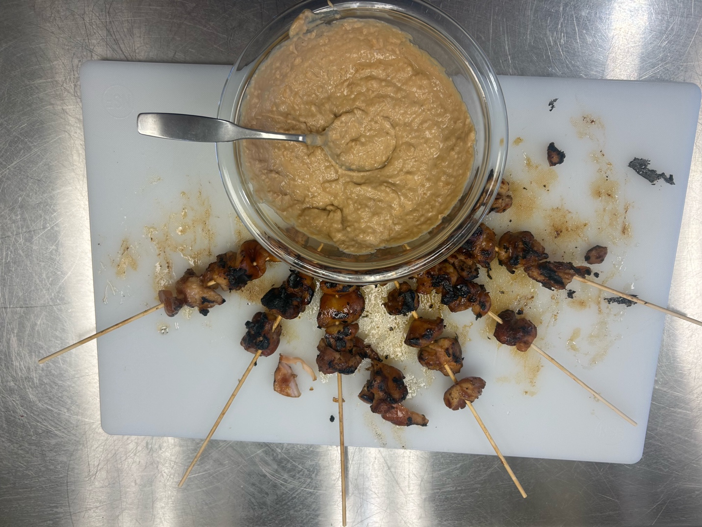 Satay chicken skewers surround a bowl of peanut sauce on a cutting board. 