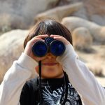 young boy peering through binoculars in a desert