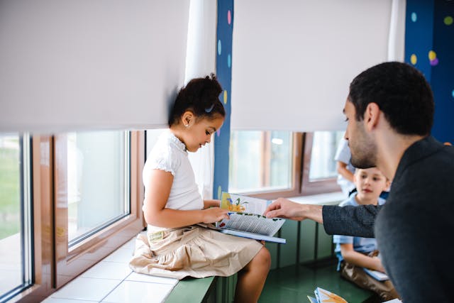 A girl sits on a wide classroom window sill. A teacher helps her find a page in a book.