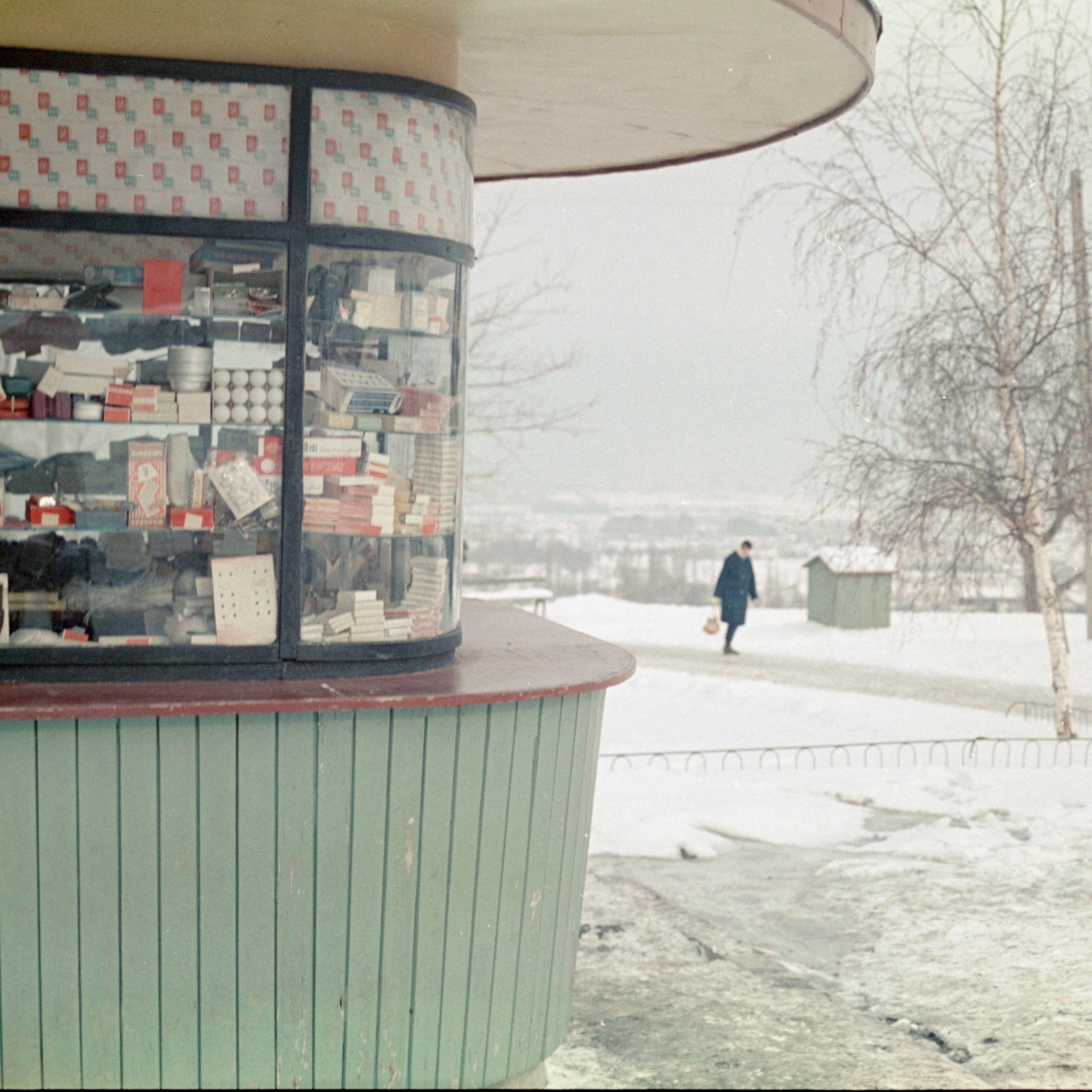 A man in a dark coat walking outside in the snow, with a shop in the foreground.