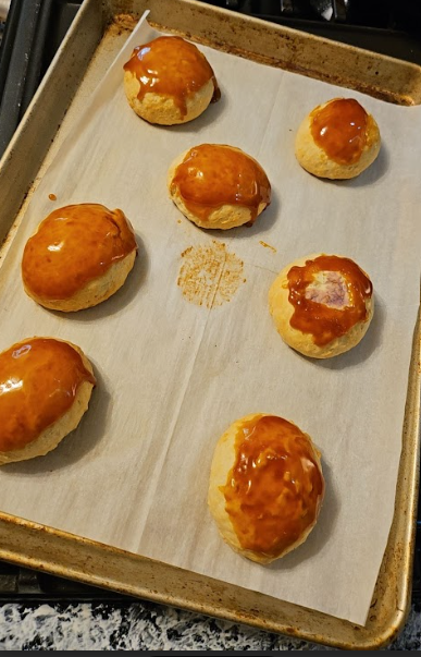 A sheet pan lined with parchment paper holds seven freshly baked Ube Bibingka Custard Buns. The buns are round, lightly golden, and topped with a shiny caramel glaze. A few have small cracks or exposed custard spots, showing natural variations from baking. The caramel appears freshly applied, giving each bun a smooth, glossy finish
