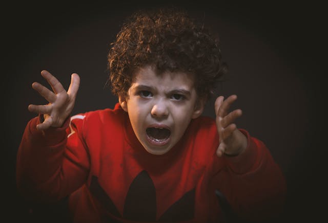 A boy in a red shirt shows frustration with his face and hands.