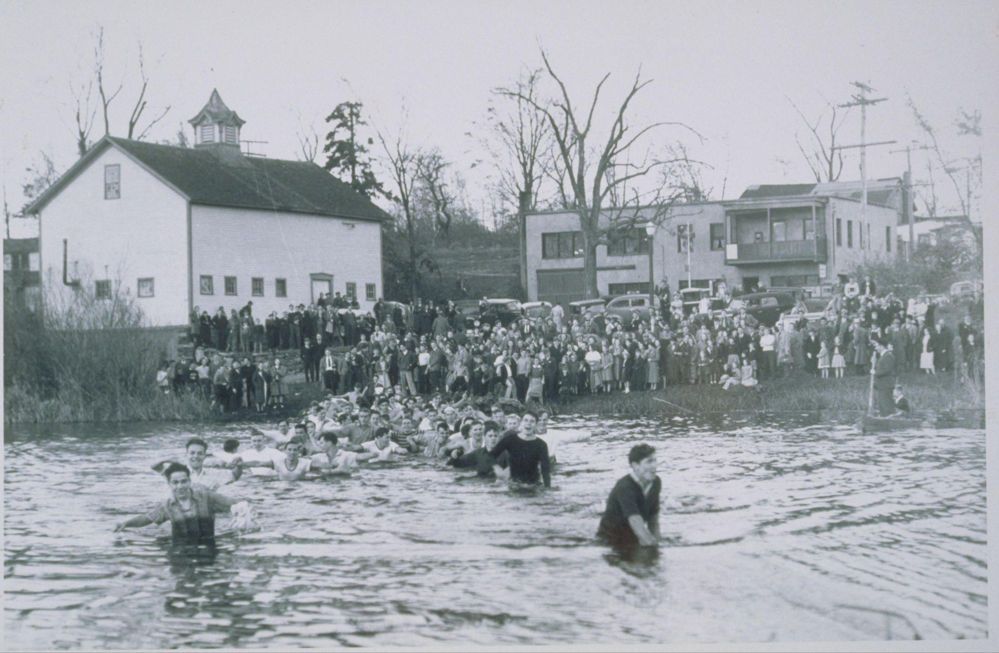 Many men wading fully-clothed and waist-deep in a body of water, with a crowd looking on.