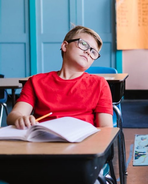 A boy in a red shirt, sitting at a desk, looking off into space.