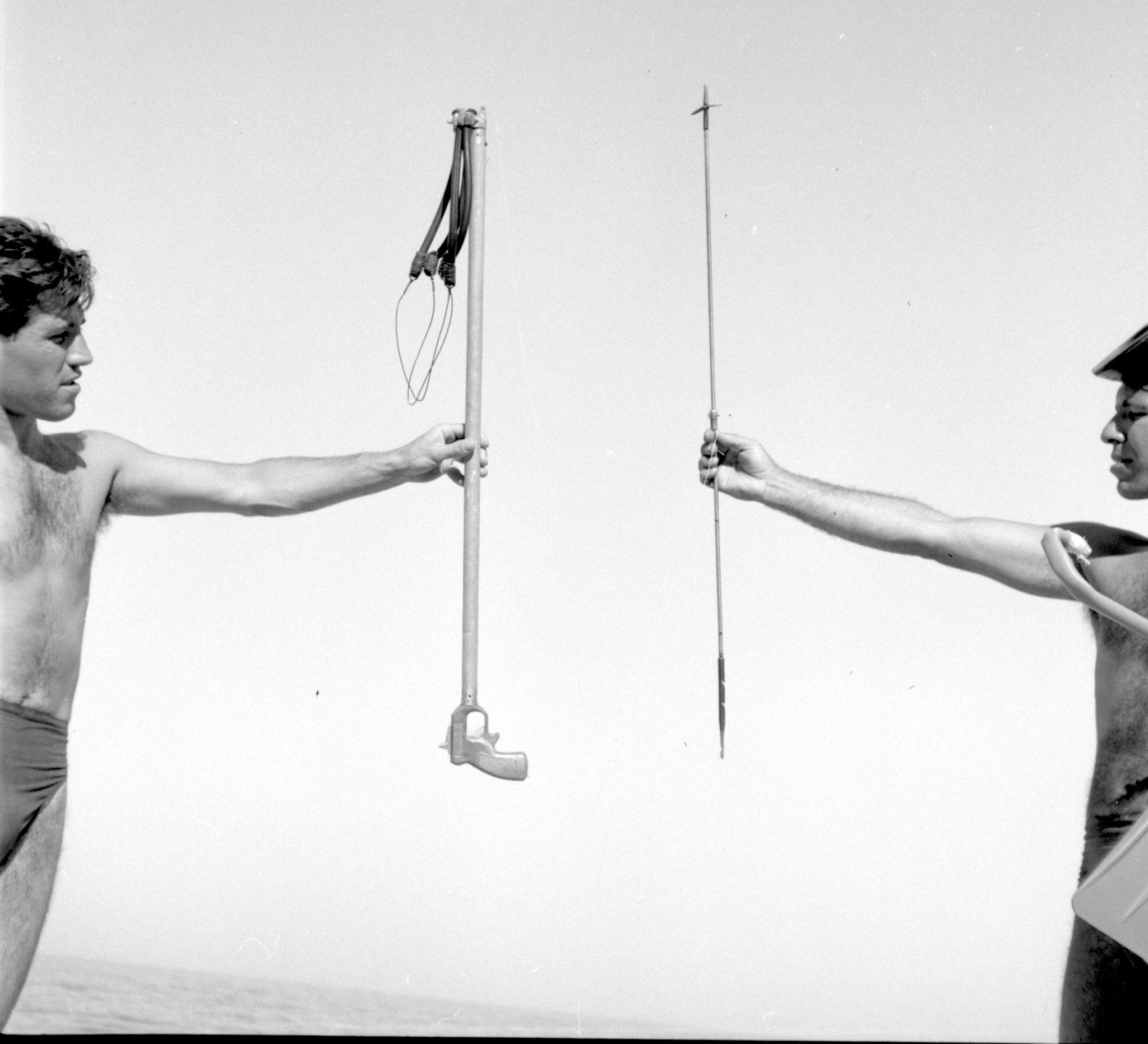 Two young men, each on the far edges of the photo and with an arm outstretched, one holding a toy rifle and another holding a spear.