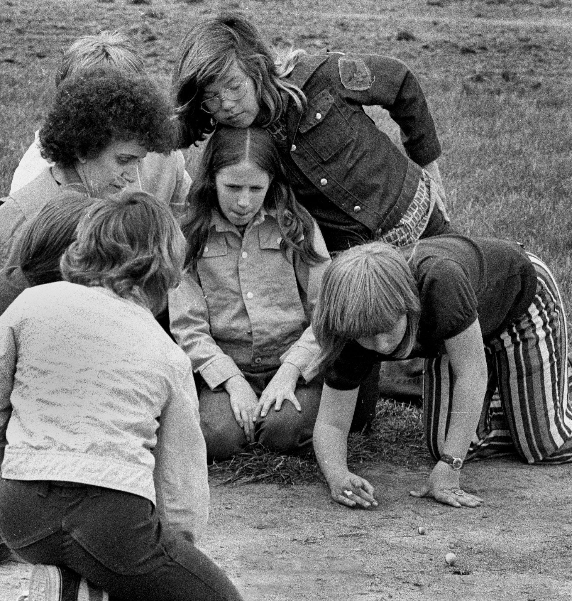 A group of seven children sit outside on the ground playing marbles. 