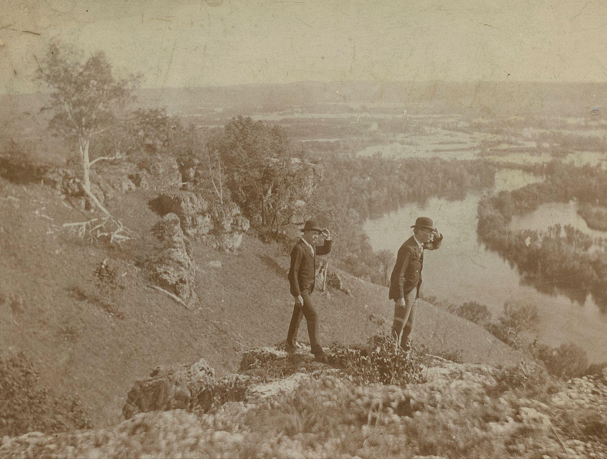 Two well-dressed men standing on a cliff look down at the river and nature below. 