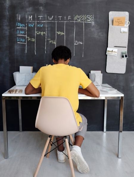 A student sits at table, facing a chalkboard. It has a weekly schedule for homework and tests.