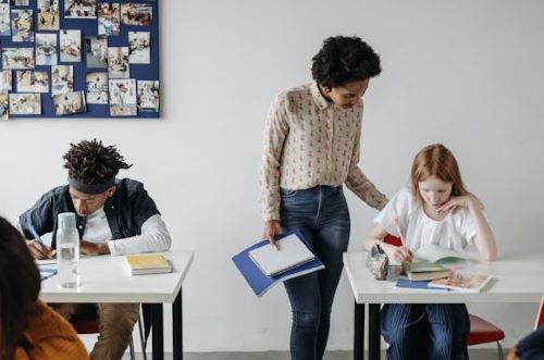Alt Text: A teacher checks on a student at a table. Another student sits on the other side of the aisle.