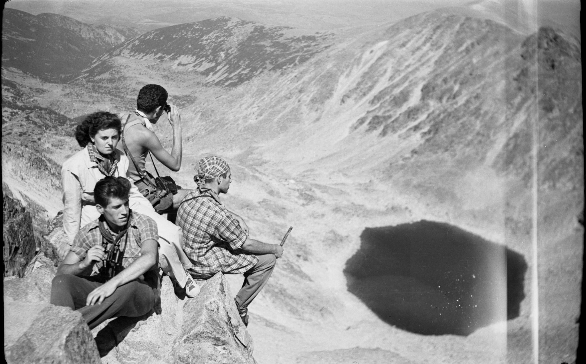 3 men and 1 woman, sitting on a rocky outcropping, overlooking a valley between 2 mountainous areas, with a lake far below them.