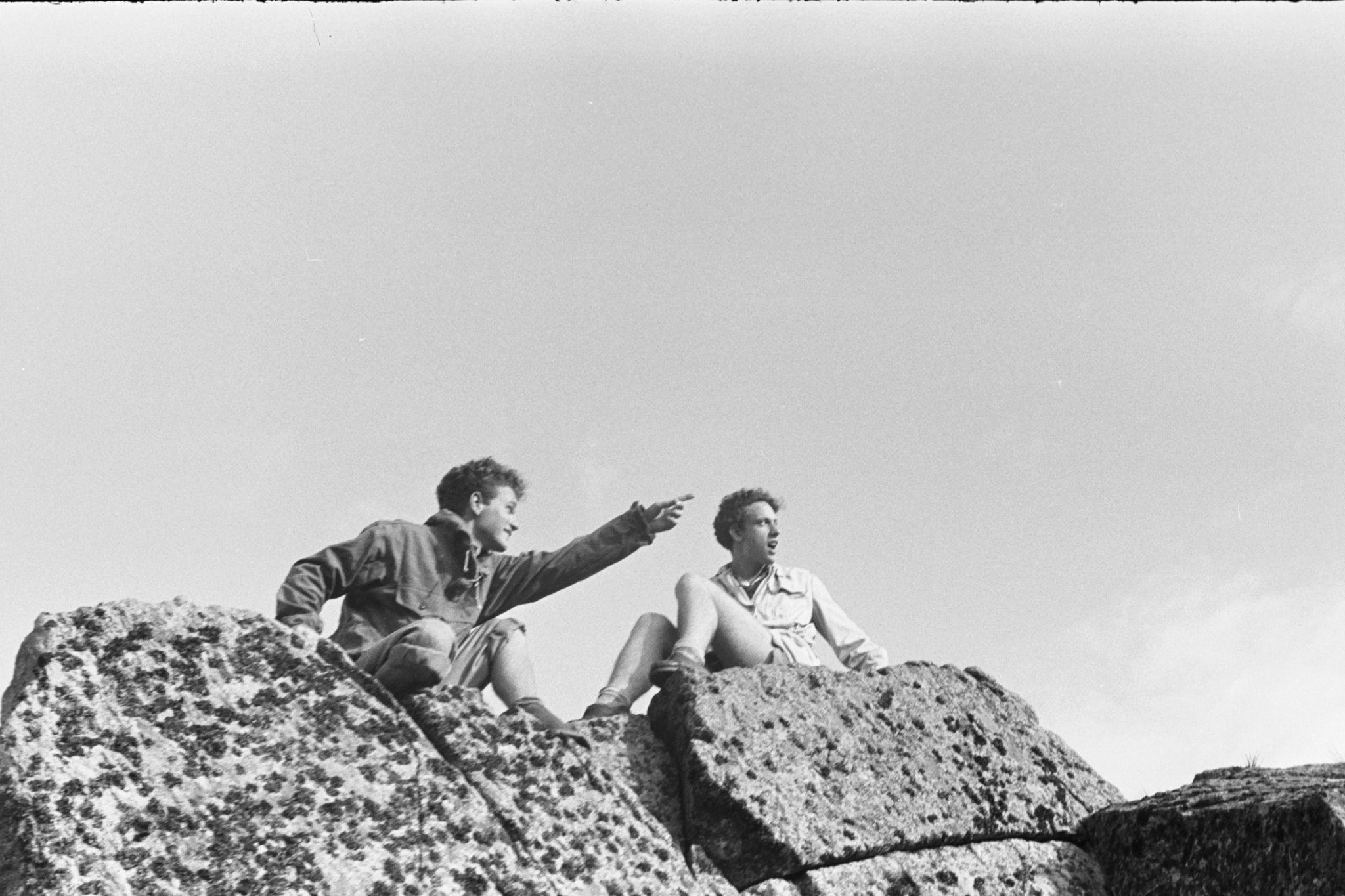 Two people sitting on rocky boulders outdoors, one pointing into the distance.